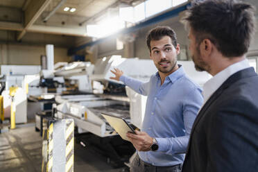 Young businessman with digital tablet gesturing toward equipment while standing by colleague at factory - DIGF12995