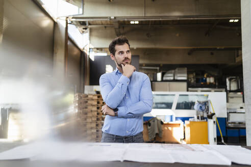 Nachdenklicher Geschäftsmann mit Hand am Kinn an einem Tisch in einer Fabrik - DIGF13008