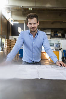 Lächelnder Mann, der sich bei der Arbeit in einer Fabrik auf einen Tisch stützt - DIGF13010