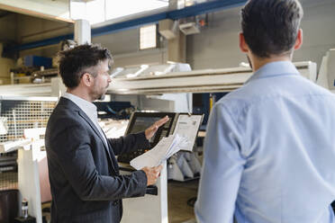 Businessman with paper having discussion with colleague while standing against machine at factory - DIGF13029