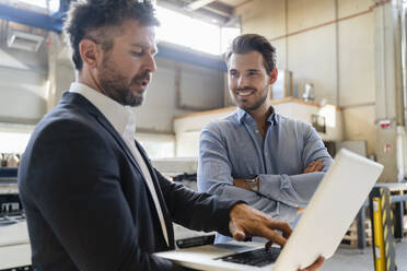 Smiling colleague standing by businessman using laptop at factory - DIGF13033