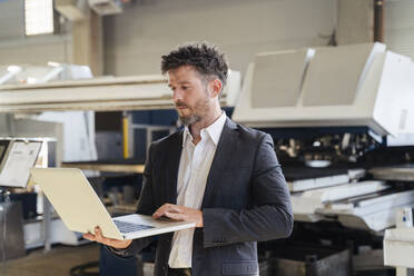 Älterer Geschäftsmann, der einen Laptop benutzt, während er in einer Fabrik steht - DIGF13034