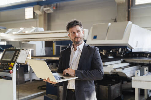 Geschäftsmann mit Laptop, der in einer Fabrik steht und wegschaut - DIGF13035