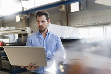 Junger Geschäftsmann, der einen Laptop benutzt, während er in einer Fabrik steht - DIGF13036