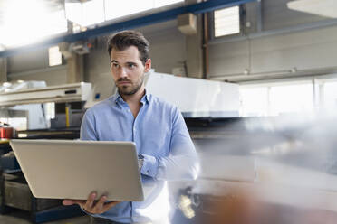 Young businessman using laptop while standing at factory - DIGF13036