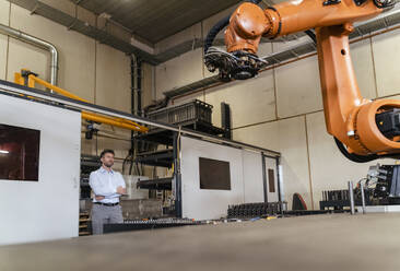 Geschäftsmann mit verschränkten Armen, der einen automatischen Roboterarm überwacht, während er in einer Fabrik steht - DIGF13097