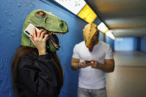 Eine Frau spricht mit einem männlichen Freund am Smartphone, während sie eine Dinosauriermaske vor einer blauen Wand trägt - IFRF00014