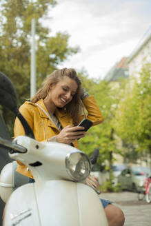 Lächelnde Frau, die auf dem Motorrad sitzend ihr Smartphone überprüft - AJOF00511