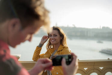 Young man taking photograph of young woman with smart phone on bridge against sky - AJOF00516