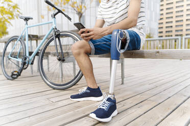 Low section of man with artificial limb using mobile phone while sitting on bench in city - JCZF00330