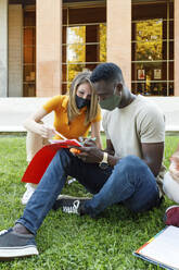 Male and female friends sitting on grass while studying together in university campus - IFRF00024
