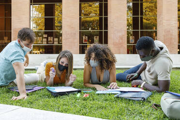 Male and female friends wearing safety mask while studying over grass in university campus - IFRF00025
