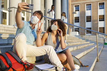 Männlicher Freund gestikuliert Frieden, während er ein Selfie mit einem Universitätsstudenten mit Schutzmaske auf einer Treppe in der Stadt macht - IFRF00043
