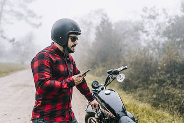 Männlicher Motorradfahrer, der über den Lautsprecher seines Smartphones spricht, während er bei nebligem Wetter neben seinem Motorrad steht - DGOF01575