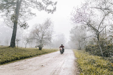 Man riding motorcycle on dirt road in foggy weather - DGOF01580