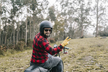 Lächelnder männlicher Biker, der auf einem Motorrad den Wald erkundet, gestikuliert - DGOF01585