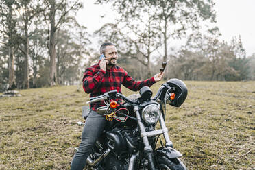 Männlicher Motorradfahrer, der auf seinem Motorrad im Wald sitzt und mit seinem Smartphone telefoniert - DGOF01589