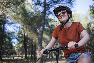 Happy young woman riding bicycle against trees at countryside - MGRF00042