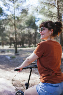 Smiling young woman riding bicycle at countryside during weekend - MGRF00044
