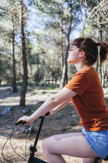 Young woman in casuals riding bicycle at countryside during weekend - MGRF00045