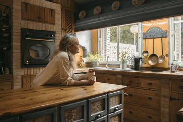 Frau mit Kaffeetasse, die wegschaut, während sie in der Küche zu Hause steht - ERRF04682