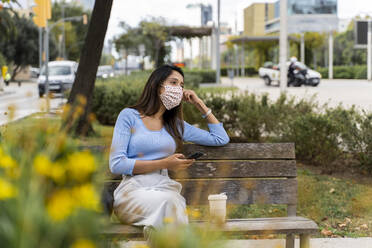 Contemplating woman looking away with smart phone while sitting on bench in city during COVID-19 - AFVF07517