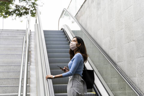 Nachdenkliche Frau, die mit ihrem Handy nach oben schaut, während sie auf der Rolltreppe steht, während COVID-19, lizenzfreies Stockfoto