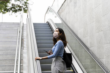 Nachdenkliche Frau, die mit ihrem Handy nach oben schaut, während sie auf der Rolltreppe steht, während COVID-19 - AFVF07525