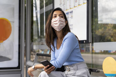 Woman with smart phone wearing face mask while waiting at bus stand during COVID-19 - AFVF07535