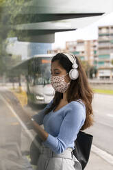 Woman in face mask wearing headphones while using smart phone at bus stand during COVID-19 - AFVF07537