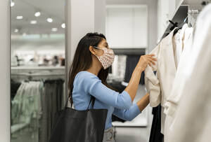 Woman wearing protective face mask while looking at suits in shopping mall - AFVF07545