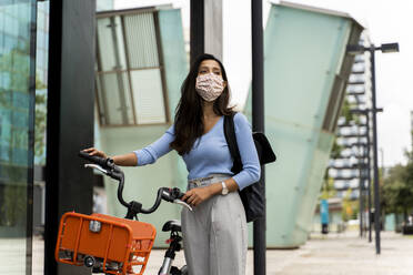 Schöne Frau mit Gesichtsmaske bei einem Spaziergang mit dem Fahrrad in der Stadt während COVID-19 - AFVF07563