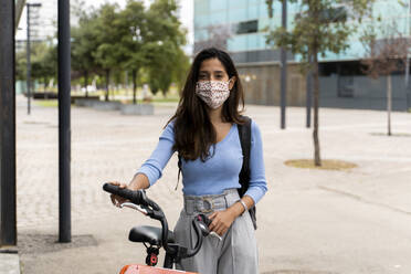 Schöne Frau mit Schutzmaske, die während der COVID-19-Pandemie mit dem Fahrrad in der Stadt steht - AFVF07564