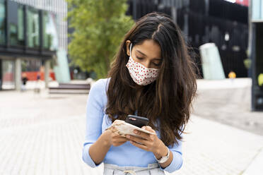 Woman wearing protective face mask while using smart phone in city during COVID-19 pandemic - AFVF07571