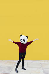Frau mit ausgestreckten Armen, die eine Panda-Maske trägt und an einer gelben Wand steht - XLGF00775