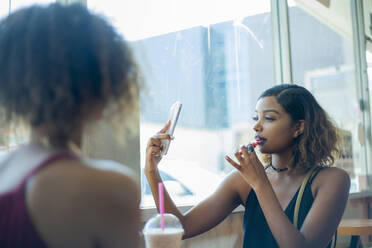 Young woman using mobile phone for applying lipstick while sitting with friend at cafe - AJOF00527