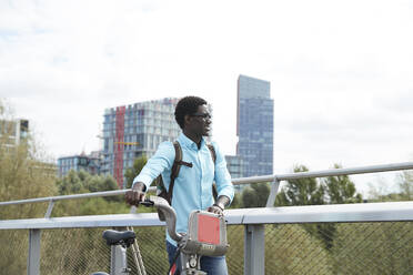 Man looking away while walking with bicycle against sky in city - PMF01518