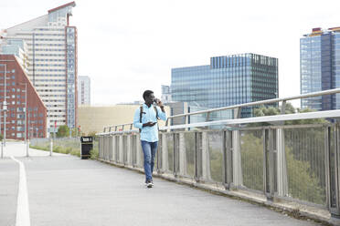 Älterer Mann trinkt Kaffee, während er sein Smartphone in der Hand hält und auf einem Fußweg in der Stadt spazieren geht - PMF01524