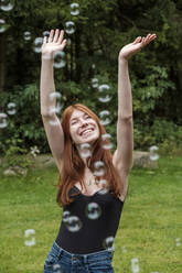 Glückliche Frau mit erhobenen Armen spielt inmitten von Seifenblasen im Garten - LBF03261