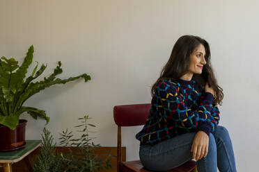 Female model looking away while sitting on chair against wall at home - VABF03877