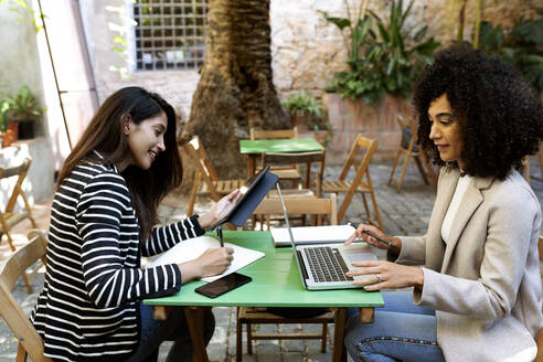 Businesswoman writing in book while female coworker using laptop sitting at cafe - VABF03891