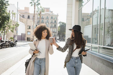 Businesswoman giving coffee cup to female colleague while walking on sidewalk in city - VABF03899