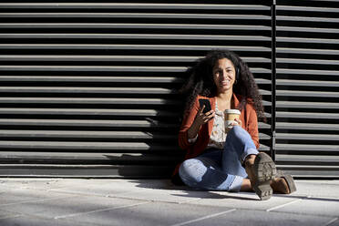 Smiling woman holding reusable cup and smart phone while sitting against black shutter - VEGF03101