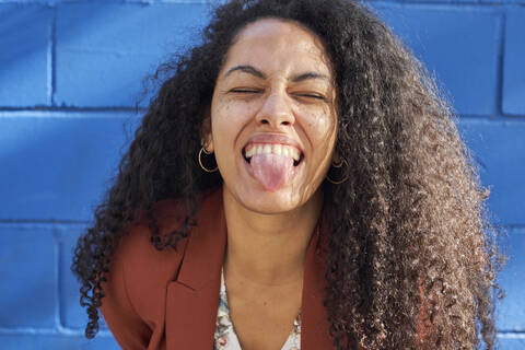 Woman sticking out tongue against blue wall on sunny day stock photo