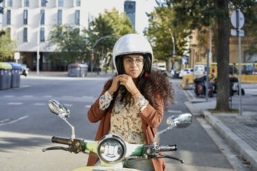 Junge Frau befestigt ihren Helm, während sie auf einem Motorroller in der Stadt sitzt - VEGF03106