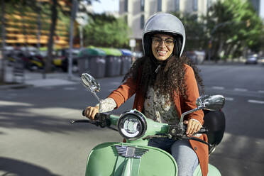 Smiling woman riding electric motor scooter in city during sunny day - VEGF03113