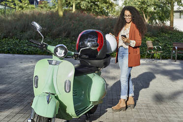 Lächelnde Frau, die einen Helm auf einem Motorroller-Container hält, während sie ein Smartphone an einem sonnigen Tag benutzt - VEGF03114