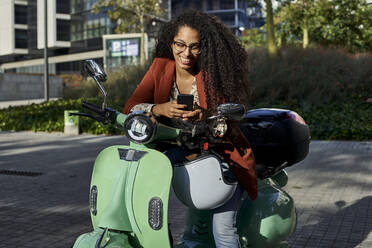 Lächelnde Frau, die ihr Smartphone benutzt, während sie auf einem Motorroller sitzt, an einem sonnigen Tag - VEGF03116