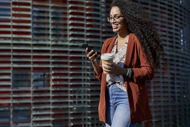 Young woman smiling using smart phone holding reusable cup while walking against building - VEGF03120