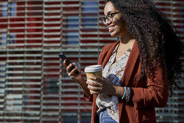 Lächelnde Frau, die ein Smartphone benutzt und einen wiederverwendbaren Becher hält, während sie vor einem Gebäude steht - VEGF03121